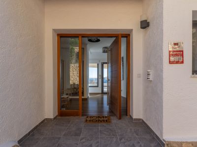 Entrada principal con puerta pivotante de madera y panel de cristal, revelando un interior luminoso con suelos de madera y vistas al mar. Muros exteriores texturizados y suelo de gres oscuro.