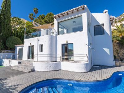 Fachada blanca de la villa con amplias ventanas y terrazas acristaladas. Destaca la piscina de mosaico azul y la tarima de composite gris, enmarcada por cipreses y palmeras.