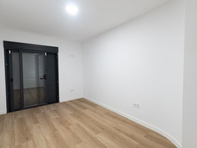Habitación luminosa con suelo laminado de madera clara y paredes blancas. Destaca una puerta corredera de cristal en gris antracita con persianas integradas que da acceso al exterior.
