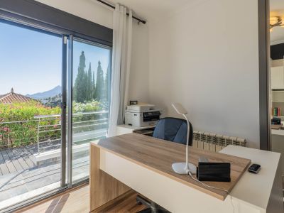 Luminoso despacho con escritorio moderno de madera, gran puerta corredera de cristal a una terraza con vegetación exuberante y vistas panorámicas a la montaña. Un espejo refleja una práctica estantería.