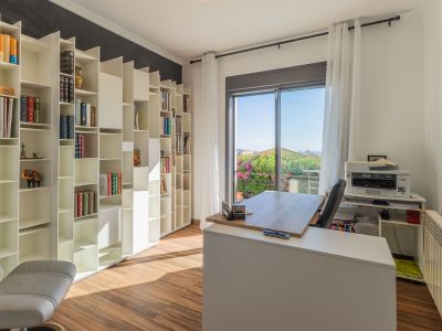 Oficina luminosa con estantería modular blanca, suelo de madera y acceso a balcón con vistas a la vegetación. El escritorio moderno se sitúa frente a la luz natural.