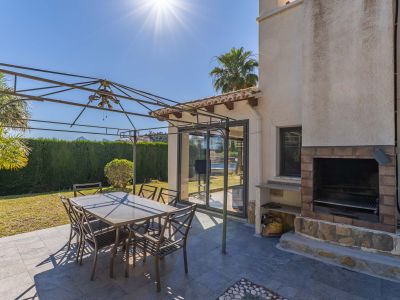 Patio exterior con suelo de gres oscuro, pérgola metálica y mesa de comedor. Incluye una barbacoa de obra con ladrillo y piedra, y acceso a través de puertas correderas de cristal.