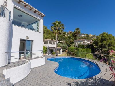 Piscina curva de mosaico azul con tarima de composite gris, adyacente a la fachada blanca con ventanal panorámico y balcón con barandilla de acero.
