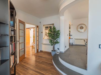 Recibidor con suelo laminado efecto madera que conecta con un espacio exterior a través de puertas acristaladas. Unas escaleras curvas de gres conducen a la zona de comedor, destacando una estantería integrada.