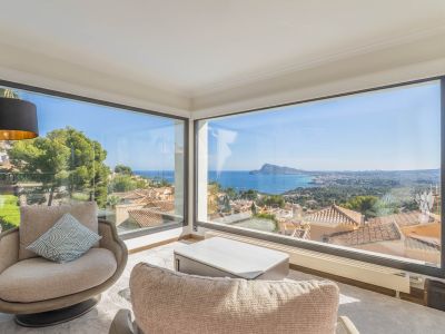 Salón con amplios ventanales de esquina que ofrecen vistas panorámicas al mar y la costa. Dos sillones y una mesa de centro moderna crean un espacio luminoso para el relax.