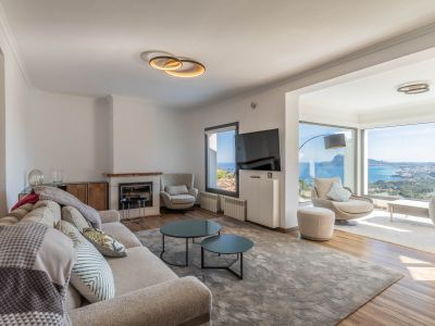 Salón moderno con sofá claro, chimenea y amplios ventanales que ofrecen vistas panorámicas al mar y la ciudad. El mobiliario contemporáneo complementa la luminosidad del espacio.