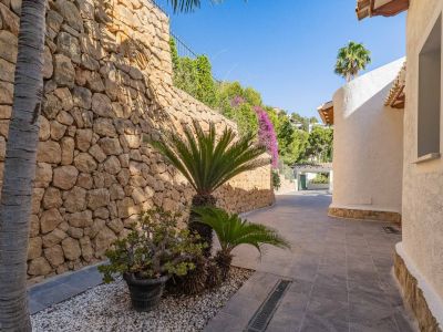 Sendero exterior con muro de piedra natural y vegetación mediterránea, flanqueado por una fachada curva de estuco blanco bajo la luz solar.
