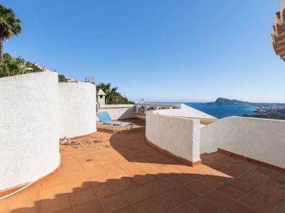 Terraza en la azotea con baldosas de terracota y muros blancos texturizados, ofreciendo vistas panorámicas al mar y la costa. Una tumbona azul invita a la relajación.