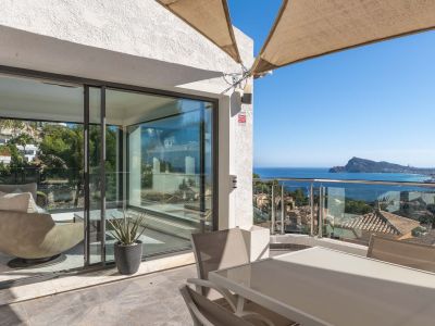 Terraza exterior con mesa de comedor y sillas, protegida por un toldo. Ofrece vistas panorámicas al mar y la costa a través de una barandilla de cristal, conectando con un interior luminoso.