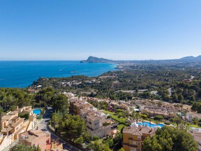 Vista aérea de la costa de Altea Hills, con el mar Mediterráneo, el Peñón de Ifach y la silueta de Benidorm al fondo. Tejados de terracota y piscinas se aprecian entre la vegetación.