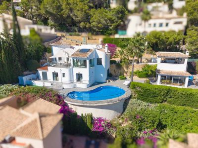 Vista aérea de la villa blanca, destacando la piscina de forma irregular con terraza de madera y barandilla. Muestra múltiples terrazas y el exuberante jardín mediterráneo.