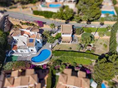 Vista aérea de la villa con su piscina de forma irregular, amplios jardines llanos con estanque y un edificio anexo. Destacan los tejados de teja y la abundante vegetación mediterránea.