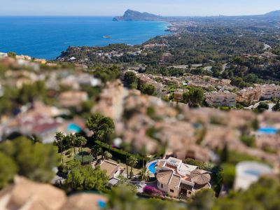 Vista aérea de una villa en Altea Hills con piscina curva, tejados de terracota y jardines exuberantes, enmarcada por el Mediterráneo y la costa.