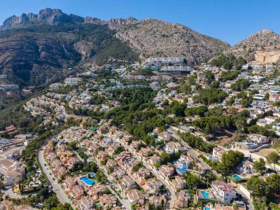 Vista aérea panorámica de Altea Hills, mostrando numerosas villas con piscinas en la ladera verde, con majestuosas montañas al fondo.