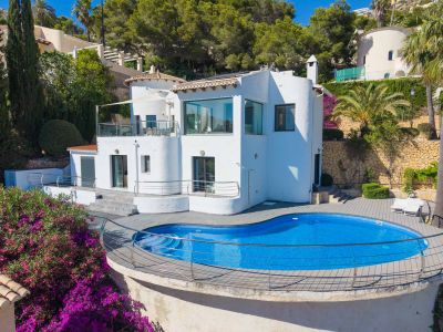 Vista exterior de la villa blanca con piscina de forma irregular y tarima de madera. Destacan la terraza superior con toldo y las vibrantes buganvillas que realzan el entorno mediterráneo.