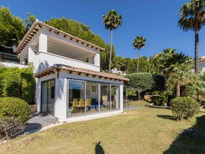 Vista exterior de la villa con fachada blanca, tejas tradicionales y amplios ventanales que conectan el interior con el jardín. Destaca el césped plano y la zona de comedor exterior bajo pérgola.