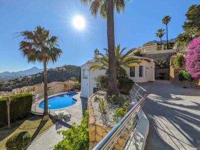 Vista exterior de la villa con piscina de forma irregular, rodeada de palmeras y un cuidado jardín. La entrada pavimentada asciende junto a muros de piedra y buganvillas, ofreciendo vistas a las montañas.