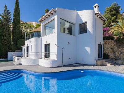 Vista exterior de la villa, destacando una piscina curva de mosaico azul y tarima gris, complementada por una ducha exterior y vegetación mediterránea.