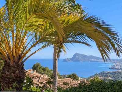 Vista panorámica al mar Mediterráneo, enmarcada por palmeras, con los tejados de Altea Hills y el Peñón de Ifach a lo lejos bajo un cielo azul.