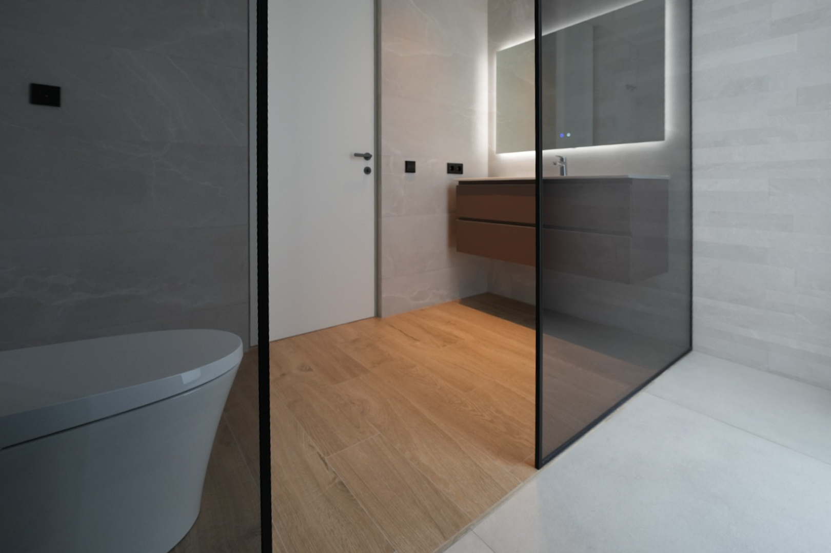 Baño de diseño moderno con inodoro suspendido, mueble de lavabo flotante de madera, espejo iluminado y ducha con mampara de cristal. Combina suelo porcelánico imitación madera y azulejos grises. Baño de diseño moderno con inodoro suspendido, mueble de lavabo flotante de madera, espejo iluminado y ducha con mampara de cristal. Combina suelo porcelánico imitación madera y azulejos grises.
