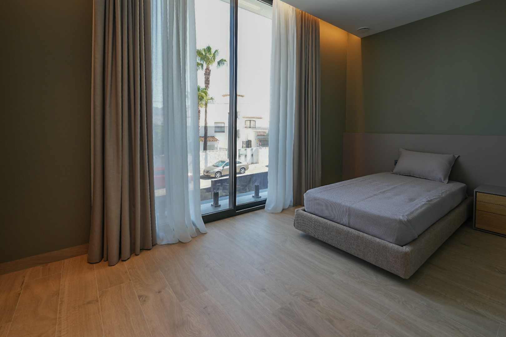Dormitorio con cama individual, suelo de madera clara y paredes verde oliva. Amplios ventanales con cortinas dobles ofrecen vistas a la calle y palmeras. Dormitorio con cama individual, suelo de madera clara y paredes verde oliva. Amplios ventanales con cortinas dobles ofrecen vistas a la calle y palmeras.
