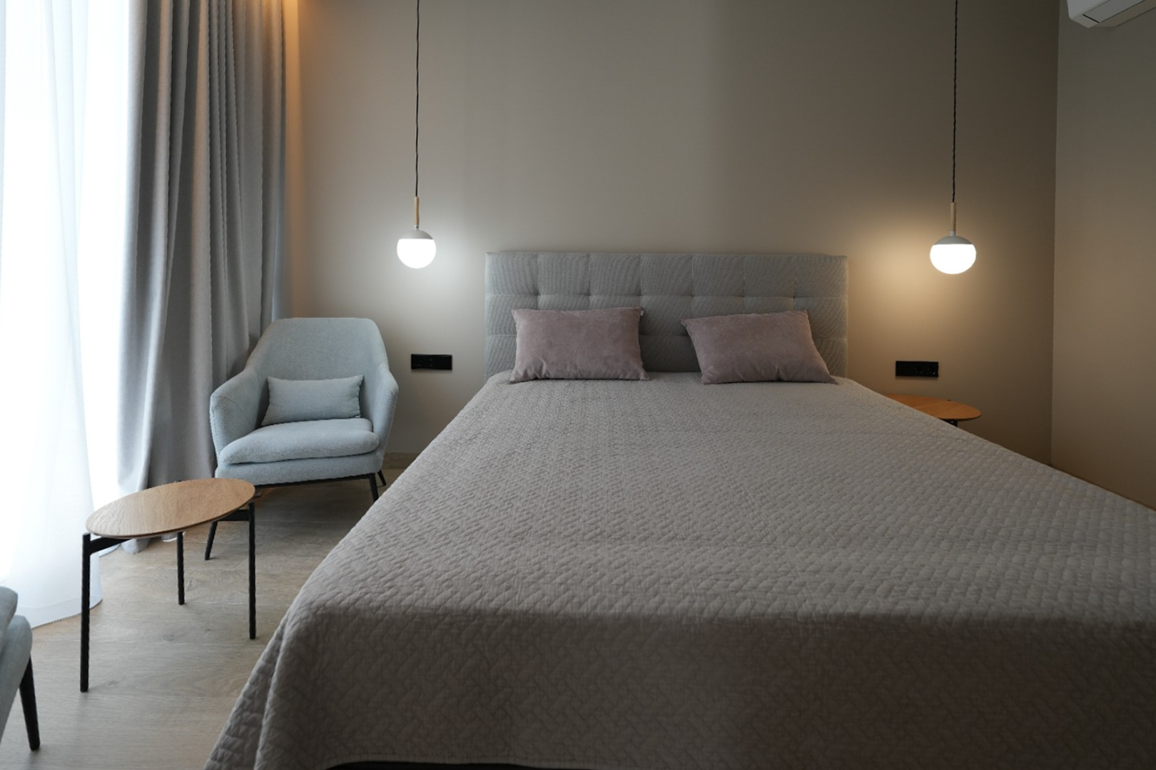 Dormitorio elegante con cama tapizada en gris, dos lámparas colgantes modernas y un sillón confortable. Cortinas claras y suelo de imitación madera crean un ambiente sereno. Dormitorio elegante con cama tapizada en gris, dos lámparas colgantes modernas y un sillón confortable. Cortinas claras y suelo de imitación madera crean un ambiente sereno.