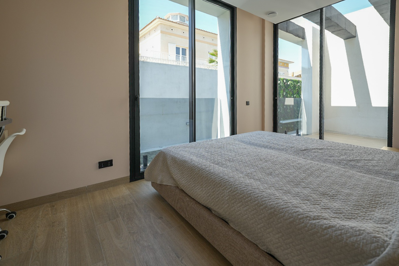 Dormitorio luminoso con suelo de madera clara y amplios ventanales correderos de perfil negro que conectan con el exterior, ofreciendo vistas a una casa vecina y vegetación. Dormitorio luminoso con suelo de madera clara y amplios ventanales correderos de perfil negro que conectan con el exterior, ofreciendo vistas a una casa vecina y vegetación.
