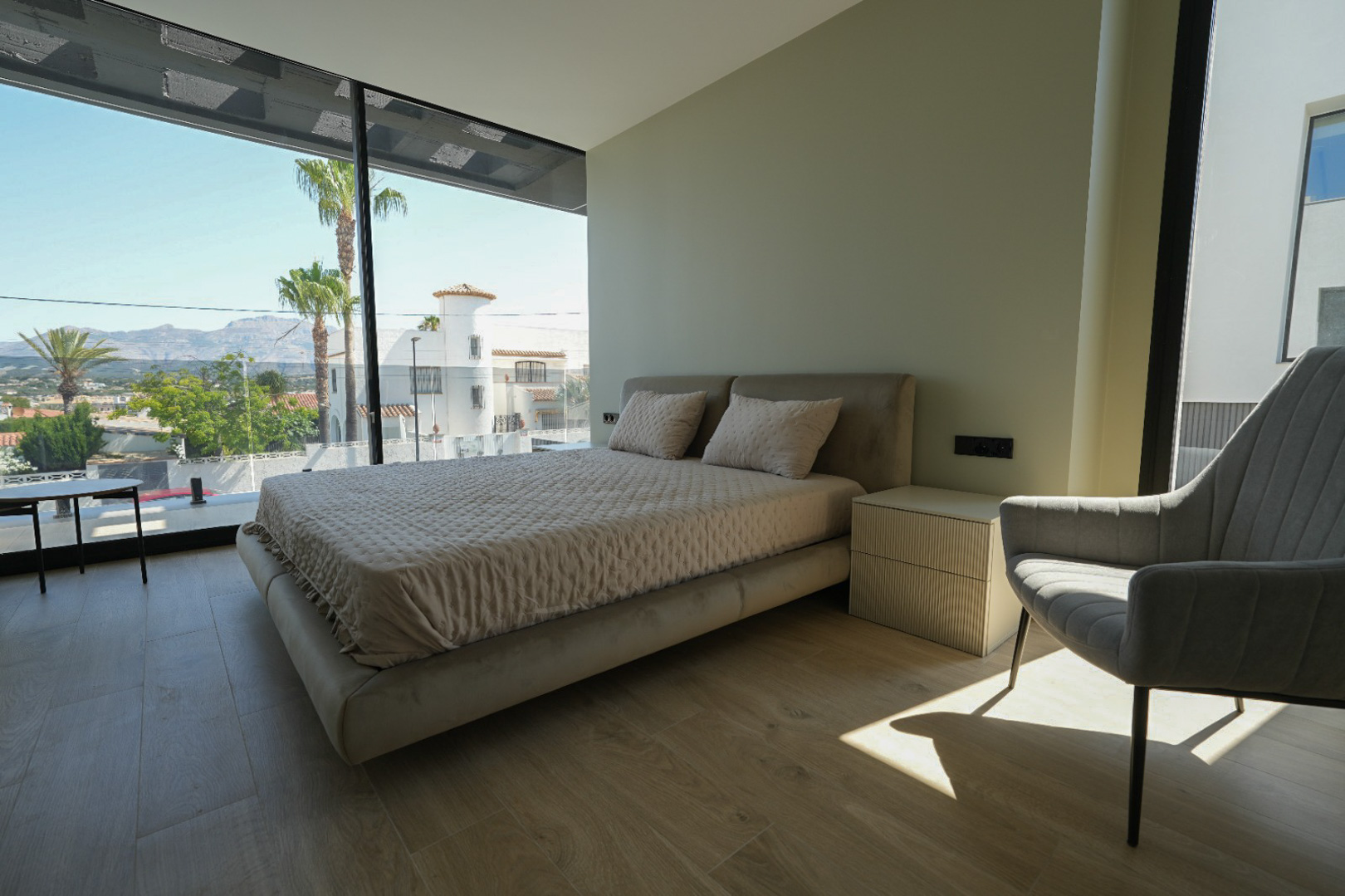 Dormitorio principal con cama tapizada, mesita de noche con textura y amplios ventanales de suelo a techo con vistas a palmeras y montañas. Dormitorio principal con cama tapizada, mesita de noche con textura y amplios ventanales de suelo a techo con vistas a palmeras y montañas.