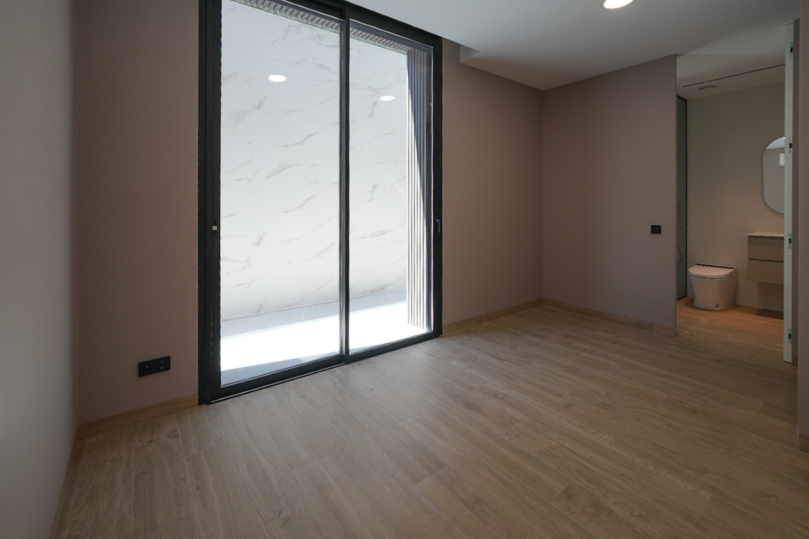 Espaciosa estancia con suelo de gres imitación madera y paredes claras. Grandes puertas correderas de cristal negro dan acceso a una terraza exterior con azulejos de mármol blanco. Baño integrado visible. Espaciosa estancia con suelo de gres imitación madera y paredes claras. Grandes puertas correderas de cristal negro dan acceso a una terraza exterior con azulejos de mármol blanco. Baño integrado visible.