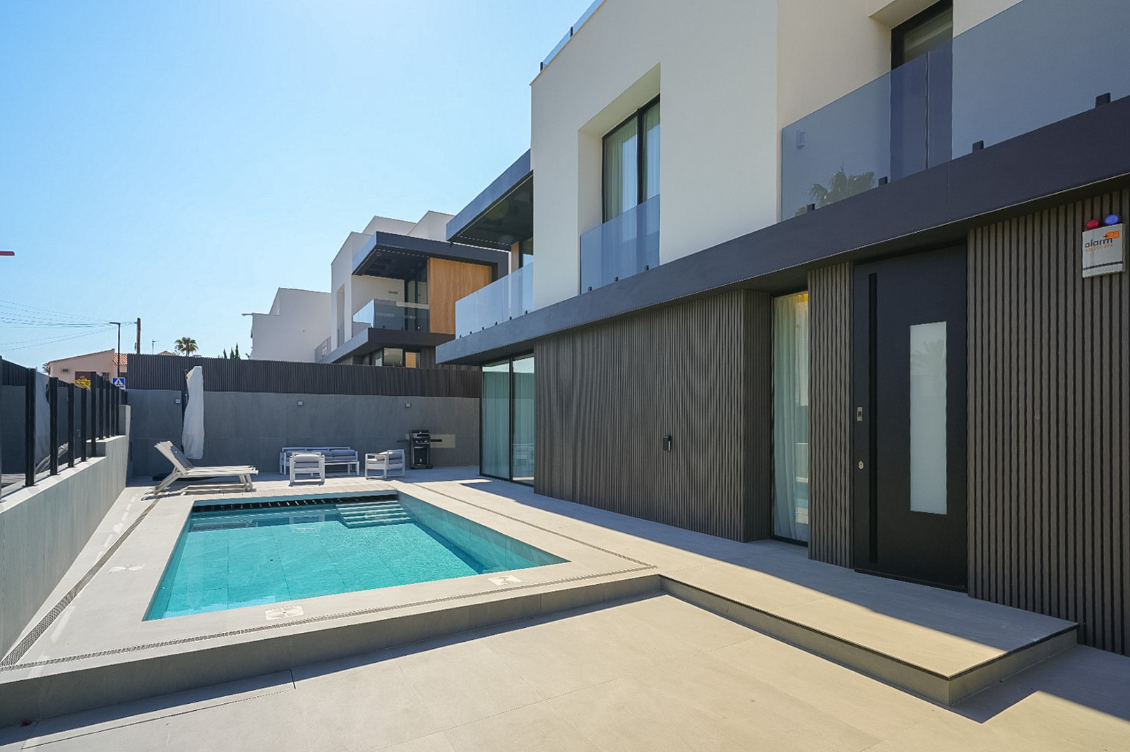 Exterior de villa moderna con piscina rectangular y escaleras integradas, rodeada de pavimento claro. Destaca la fachada inferior con lamas oscuras y la puerta de entrada principal. Exterior de villa moderna con piscina rectangular y escaleras integradas, rodeada de pavimento claro. Destaca la fachada inferior con lamas oscuras y la puerta de entrada principal.