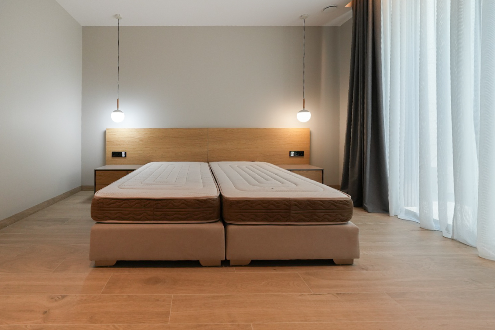 Habitación con cama doble de bases tapizadas, cabecero de madera clara con mesitas de noche integradas y dos lámparas colgantes modernas. Ventanal con cortinas claras. Habitación con cama doble de bases tapizadas, cabecero de madera clara con mesitas de noche integradas y dos lámparas colgantes modernas. Ventanal con cortinas claras.