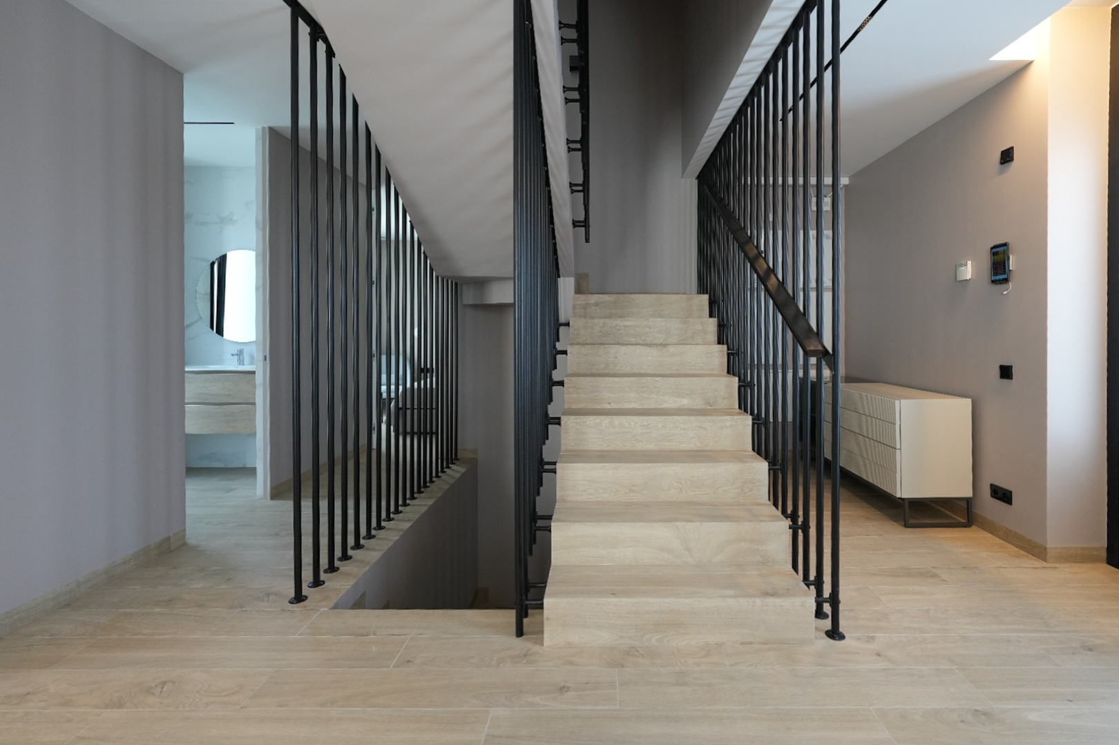 Interior moderno con escalera de peldaños en madera clara y barandilla vertical negra. Panel de control domótico en pared, con acceso a baño contemporáneo con espejo redondo. Interior moderno con escalera de peldaños en madera clara y barandilla vertical negra. Panel de control domótico en pared, con acceso a baño contemporáneo con espejo redondo.
