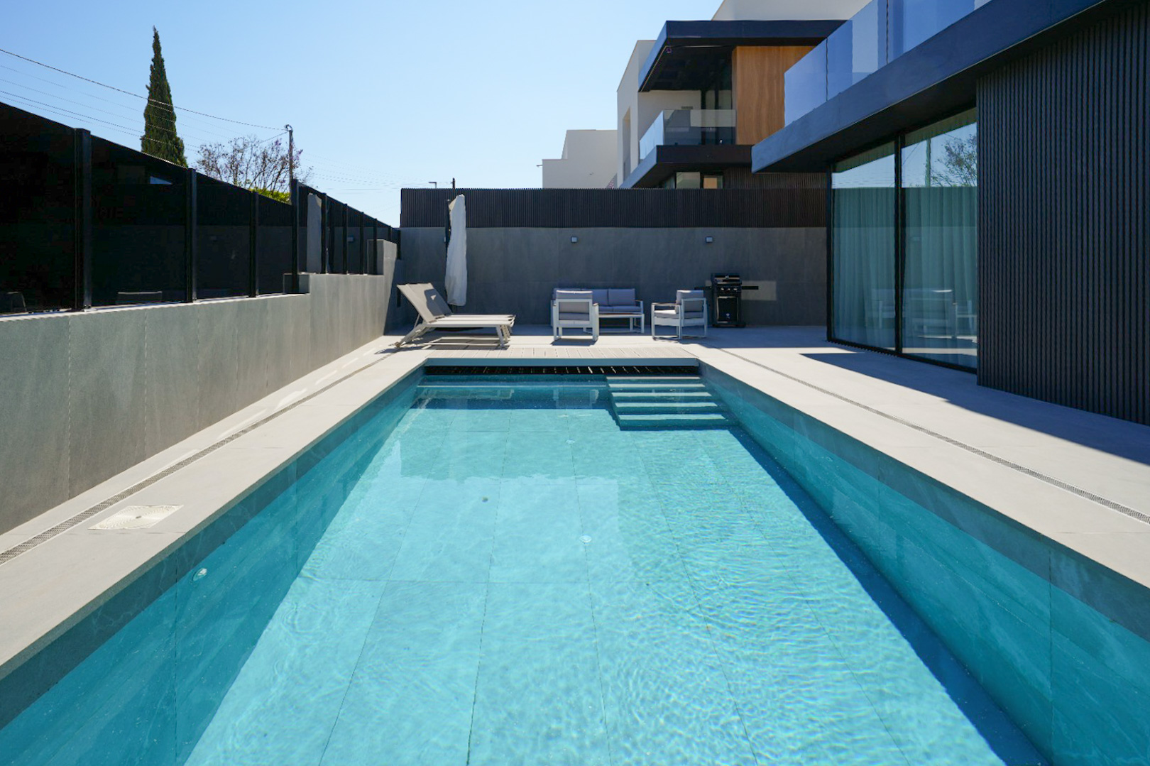 La zona exterior presenta una piscina rectangular con azulejos celestes y escaleras, rodeada de pavimento gris claro. Un moderno conjunto de salón y tumbona invitan a la relajación junto a la elegante fachada. La zona exterior presenta una piscina rectangular con azulejos celestes y escaleras, rodeada de pavimento gris claro. Un moderno conjunto de salón y tumbona invitan a la relajación junto a la elegante fachada.