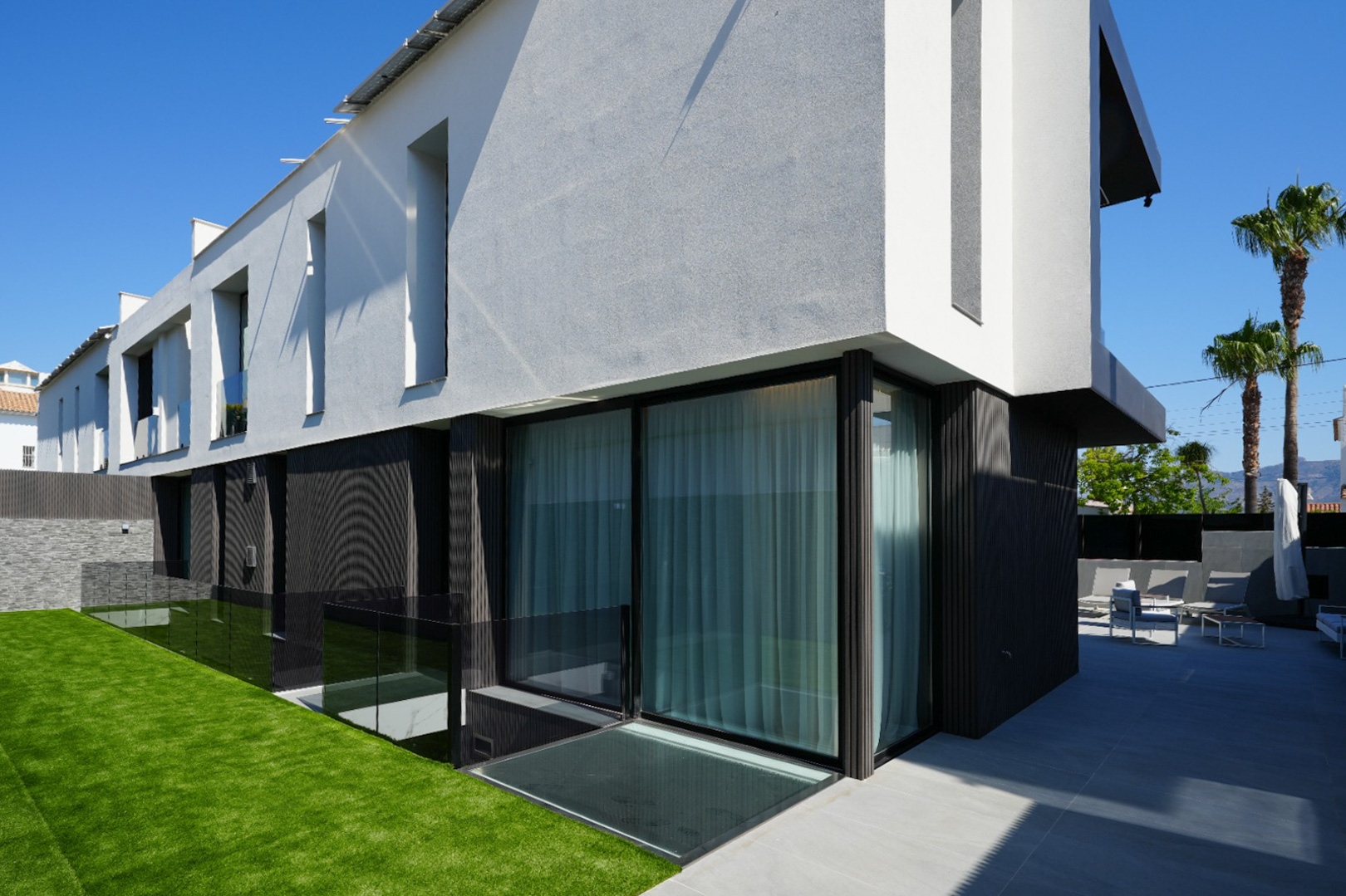 Moderna fachada exterior con contraste de texturas en blanco y gris, grandes ventanales, jardín de césped artificial y patio con mobiliario exterior. Moderna fachada exterior con contraste de texturas en blanco y gris, grandes ventanales, jardín de césped artificial y patio con mobiliario exterior.