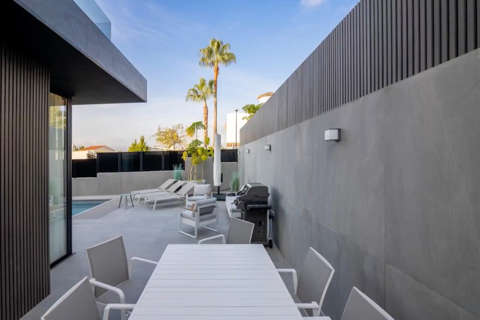 Moderna terraza exterior con mesa de comedor y sillas blancas, zona lounge y barbacoa, enmarcada por paredes de gres gris y celosías oscuras, con vista a la piscina. Moderna terraza exterior con mesa de comedor y sillas blancas, zona lounge y barbacoa, enmarcada por paredes de gres gris y celosías oscuras, con vista a la piscina.