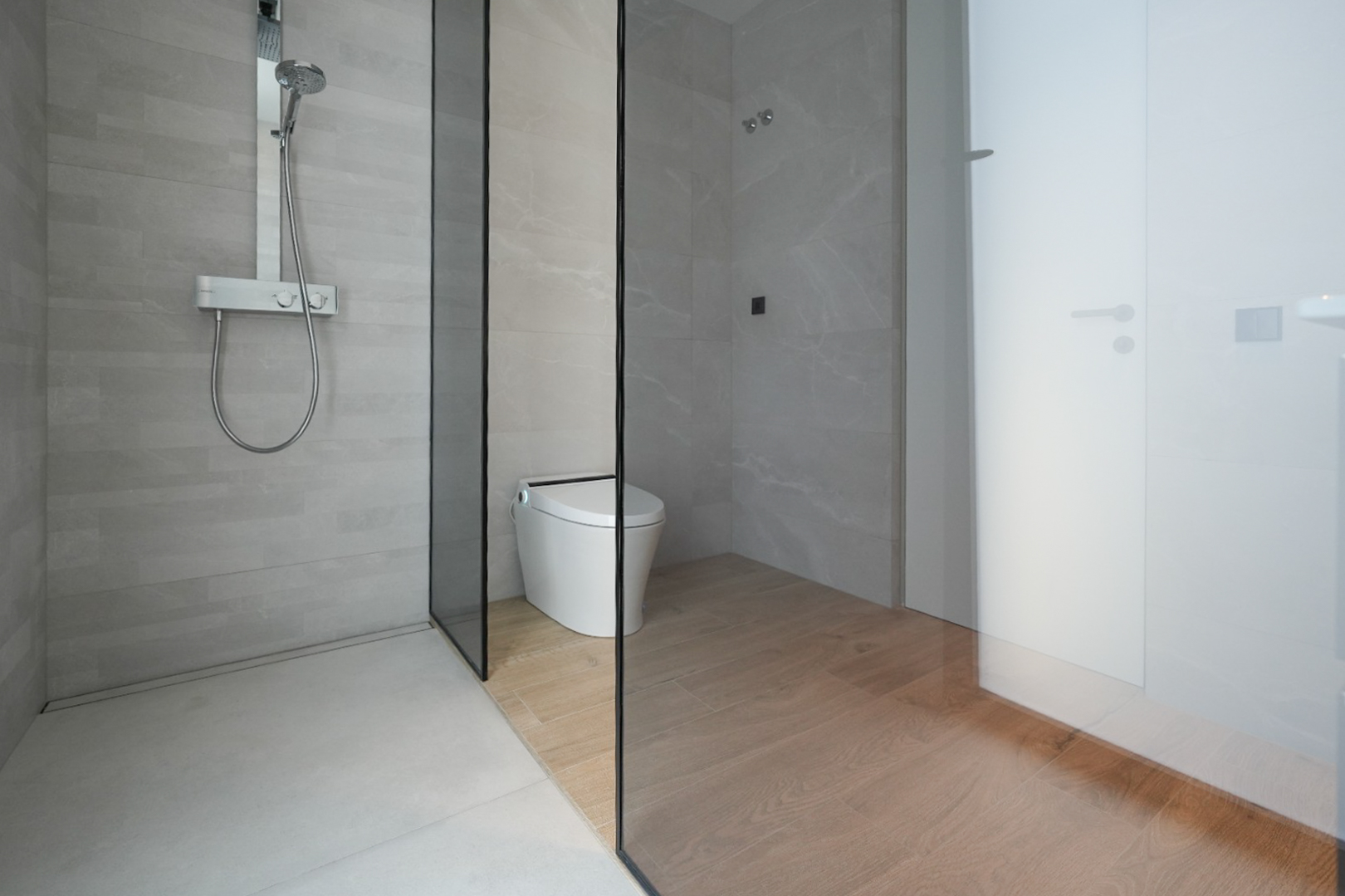 Moderno baño con ducha a ras de suelo, azulejos grises claros y grifería termostática cromada. Una mampara de cristal separa la ducha del inodoro inteligente con suelo de baldosas imitación madera. Moderno baño con ducha a ras de suelo, azulejos grises claros y grifería termostática cromada. Una mampara de cristal separa la ducha del inodoro inteligente con suelo de baldosas imitación madera.
