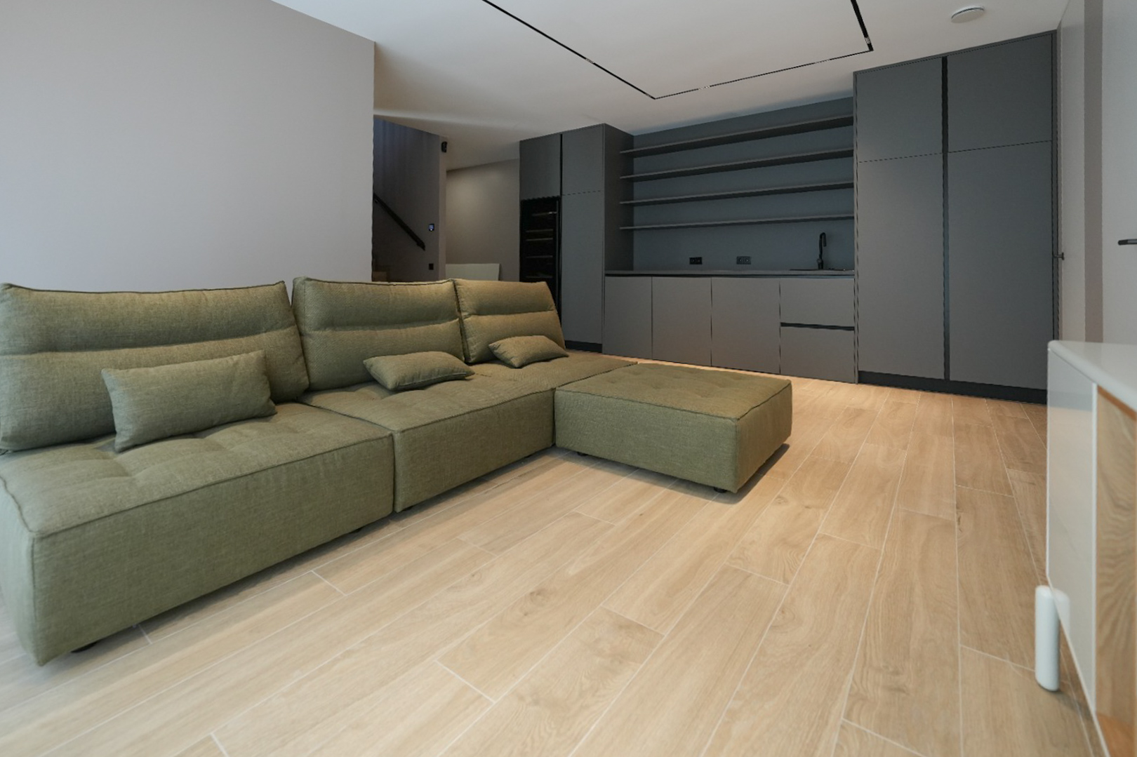 Sofá modular verde oliva en sala de estar de concepto abierto con suelo de baldosas imitación madera. Al fondo, mobiliario de cocina oscuro con fregadero y estantes. Sofá modular verde oliva en sala de estar de concepto abierto con suelo de baldosas imitación madera. Al fondo, mobiliario de cocina oscuro con fregadero y estantes.