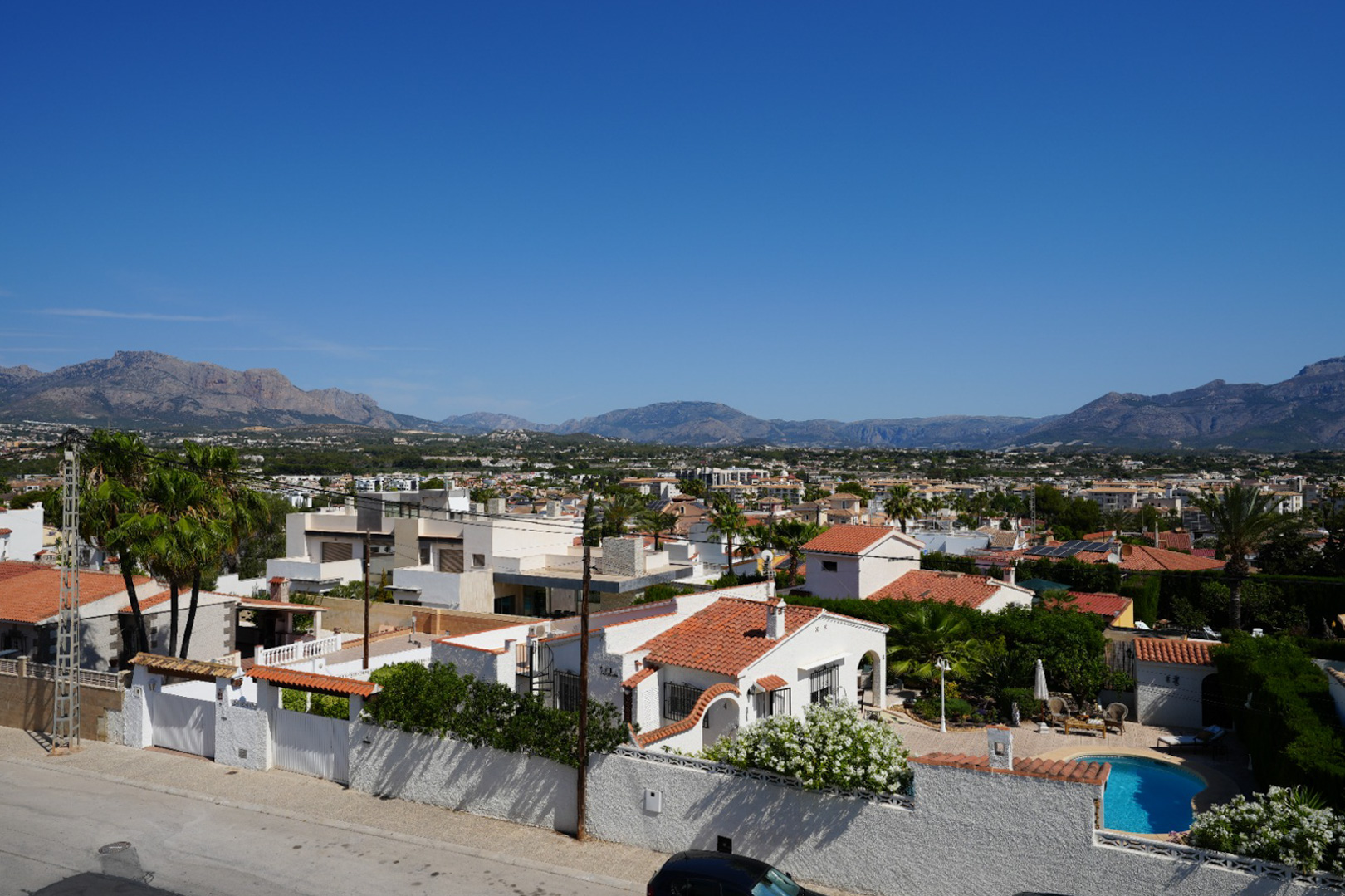 Vista elevada del barrio residencial de Albir, mostrando diversidad arquitectónica, exuberante vegetación y una piscina privada, con la imponente sierra al fondo bajo un cielo despejado. Vista elevada del barrio residencial de Albir, mostrando diversidad arquitectónica, exuberante vegetación y una piscina privada, con la imponente sierra al fondo bajo un cielo despejado.