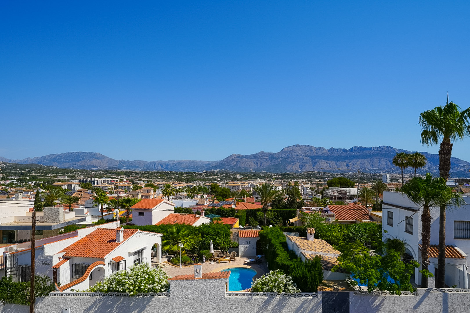 Vista elevada del entorno residencial de Albir, destacando una piscina privada, casas con tejados de teja y exuberante vegetación mediterránea, con montañas al fondo. Vista elevada del entorno residencial de Albir, destacando una piscina privada, casas con tejados de teja y exuberante vegetación mediterránea, con montañas al fondo.