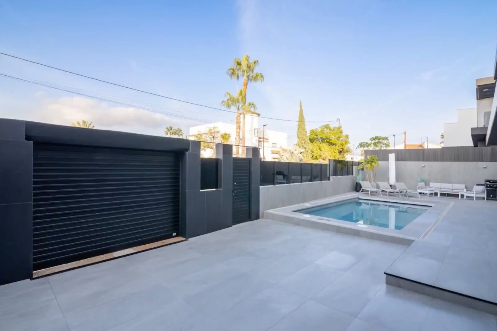 Vista exterior con piscina privada, patio de baldosas claras y puerta de garaje oscura con acceso peatonal seguro. Vista exterior con piscina privada, patio de baldosas claras y puerta de garaje oscura con acceso peatonal seguro.