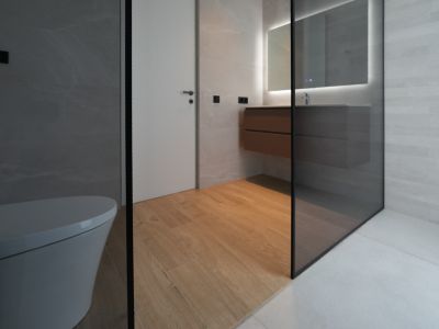 Baño de diseño moderno con inodoro suspendido, mueble de lavabo flotante de madera, espejo iluminado y ducha con mampara de cristal. Combina suelo porcelánico imitación madera y azulejos grises.
