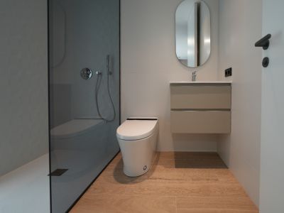 Baño moderno con ducha a ras de suelo de cristal oscuro y pared texturizada, inodoro suspendido, mueble de lavabo flotante y suelo de gres porcelánico efecto madera.