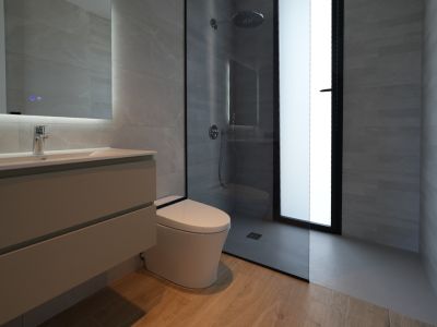 Baño moderno con lavabo flotante, espejo iluminado, inodoro suspendido y ducha a ras de suelo con rociador tipo lluvia, revestido con azulejos grises y suelo de efecto madera.