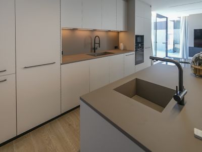 Cocina de diseño con isla central y doble fregadero integrado. Armarios minimalistas en tonos neutros, electrodomésticos empotrados y suelo de madera clara, con acceso directo al exterior.