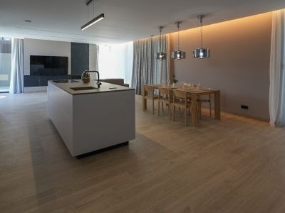 Cocina de diseño con isla central y electrodomésticos integrados, conectada a un comedor con mesa de madera y lámparas colgantes modernas. Suelo de porcelánico efecto madera.