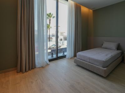 Dormitorio con cama individual, suelo de madera clara y paredes verde oliva. Amplios ventanales con cortinas dobles ofrecen vistas a la calle y palmeras.