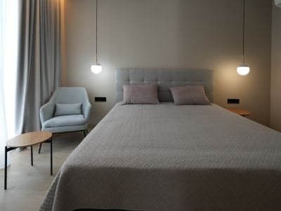 Dormitorio elegante con cama tapizada en gris, dos lámparas colgantes modernas y un sillón confortable. Cortinas claras y suelo de imitación madera crean un ambiente sereno.