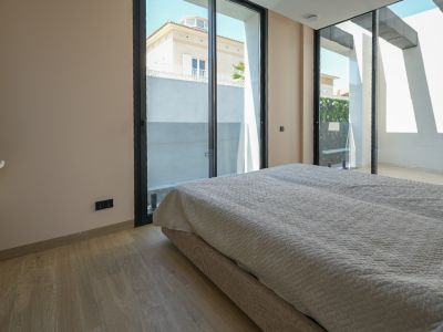 Dormitorio luminoso con suelo de madera clara y amplios ventanales correderos de perfil negro que conectan con el exterior, ofreciendo vistas a una casa vecina y vegetación.