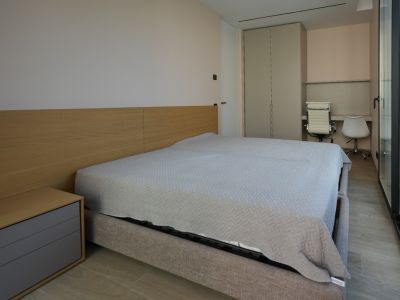 Dormitorio moderno con cama tapizada gris claro, cabecero de madera y mesita integrada. Dispone de armario empotrado, zona de escritorio doble y amplios ventanales.