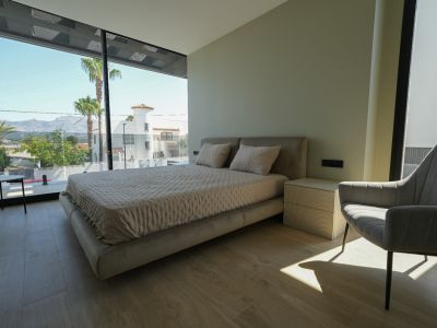 Dormitorio principal con cama tapizada, mesita de noche con textura y amplios ventanales de suelo a techo con vistas a palmeras y montañas.
